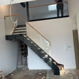 Radosław Piłat Spaw-Pil - Montaż nowoczesnych schodów z czarną konstrukcją i szklanymi balustradami w trakcie prac wykończeniowych. Widoczne kable, drabina i folie ochronne na podłodze.