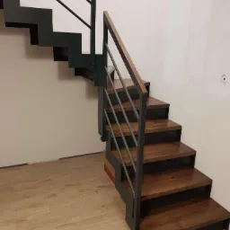 Nowoczesne schody z drewnianymi stopniami i metalową balustradą, widoczne od dołu, z drewnianą poręczą, na tle białej ściany i podłogi z płytek imitujących drewno.