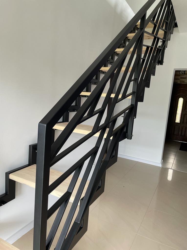 Nowoczesne schody z drewnianymi stopniami i czarną, geometryczną balustradą metalową, widok z dołu, jasna podłoga i drzwi w tle.