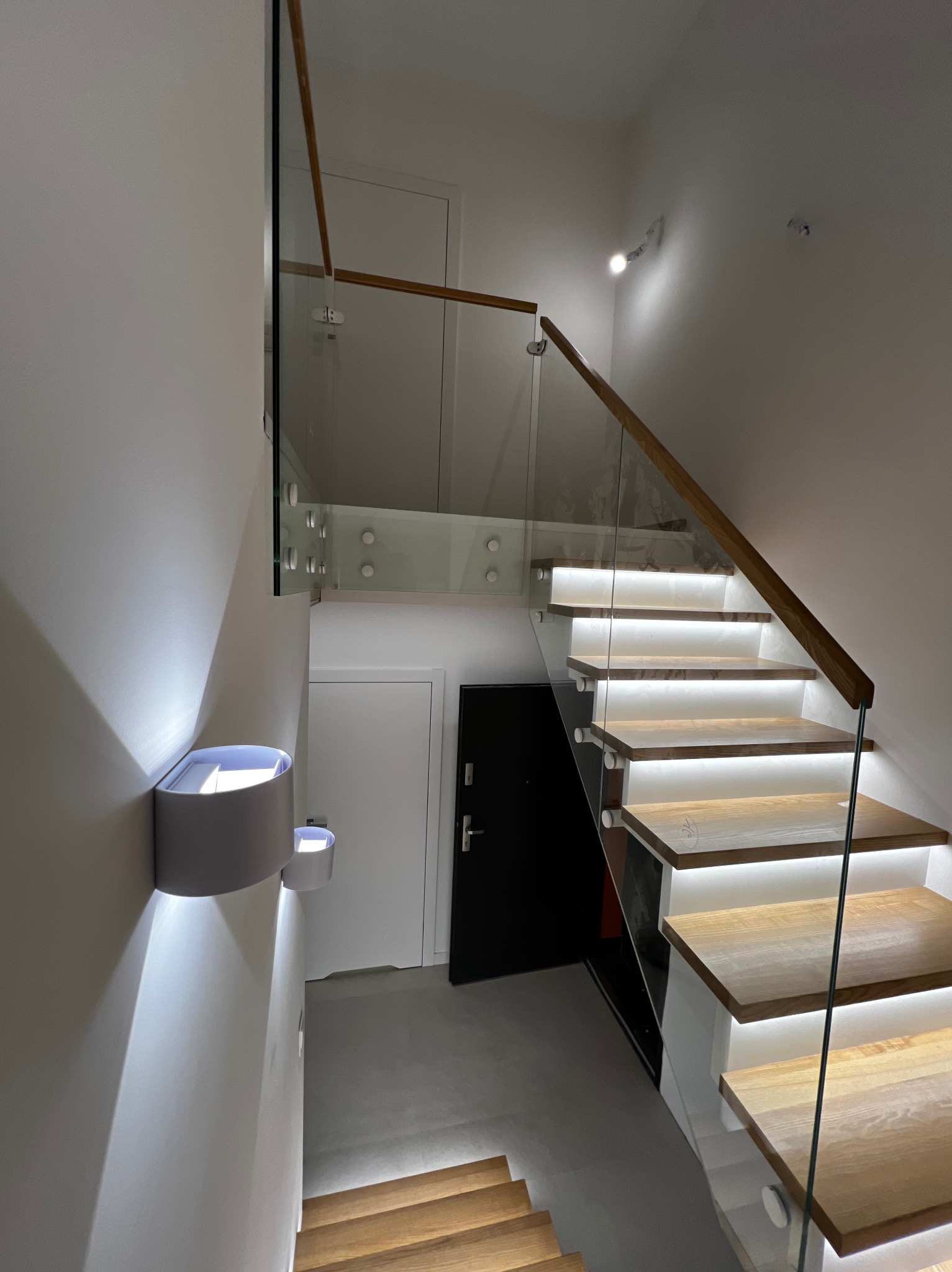 Nowoczesne schody z drewnianymi stopniami podświetlonymi LED, balustrada szklana z drewnianą poręczą, oświetlenie ścienne w kształcie tuby.