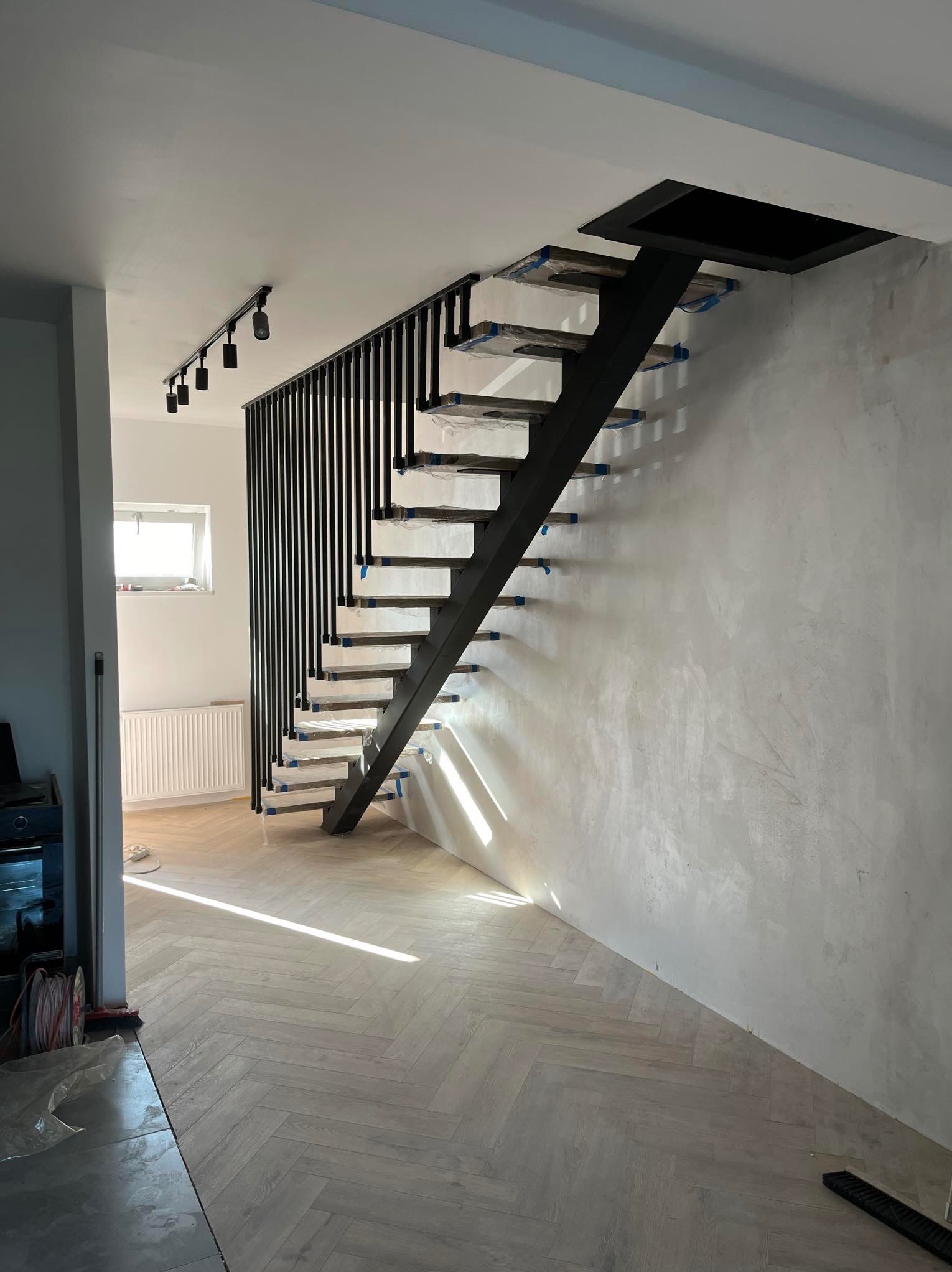 Nowoczesne schody z czarną metalową konstrukcją i drewnianymi stopniami, zabezpieczonymi folią, w jasnym wnętrzu z parkietem ułożonym w jodełkę.