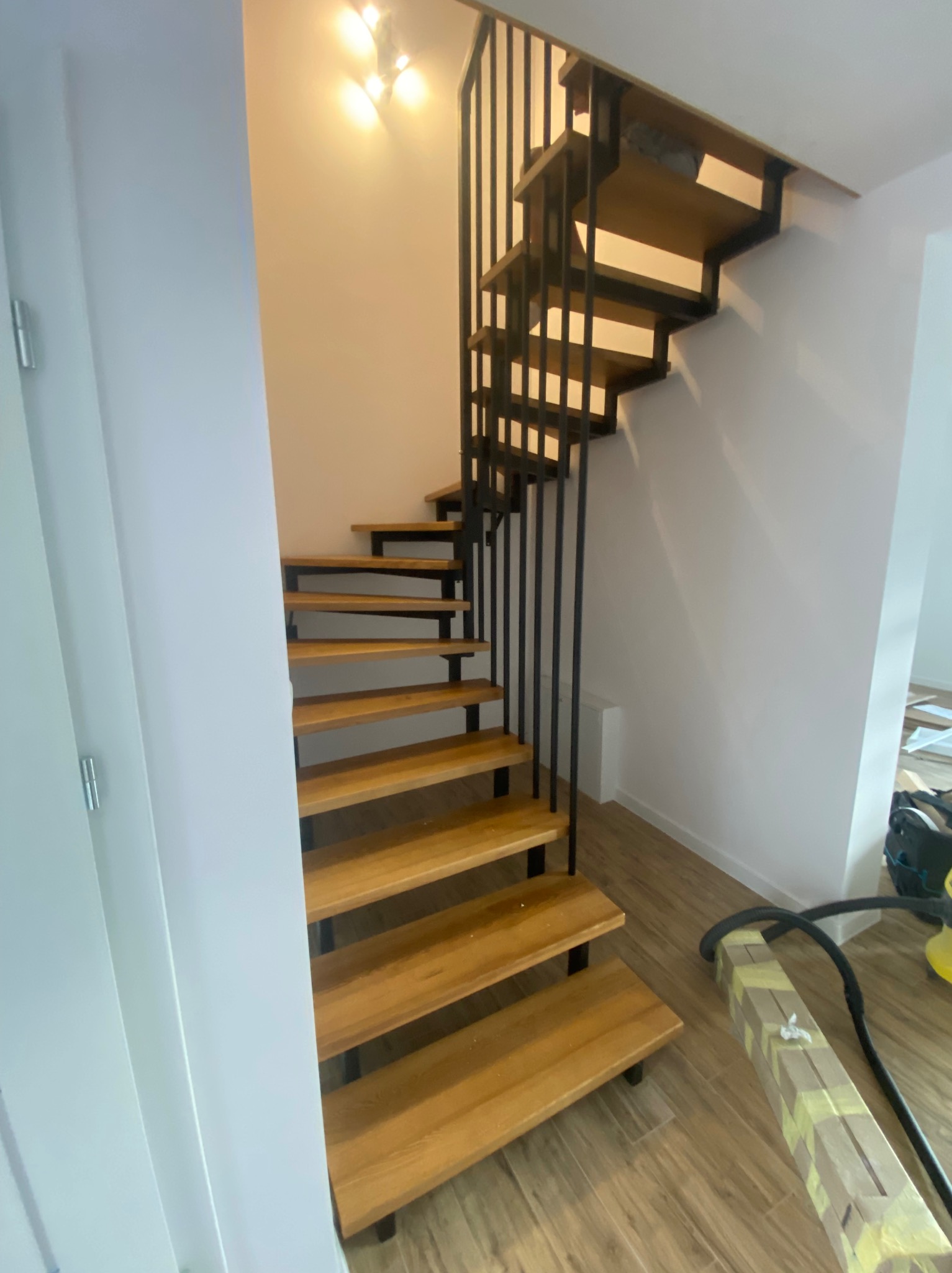 Ażurowe schody z drewnianymi stopniami i czarną metalową konstrukcją, widoczne z dołu, z elementami wykończeniowymi na podłodze.