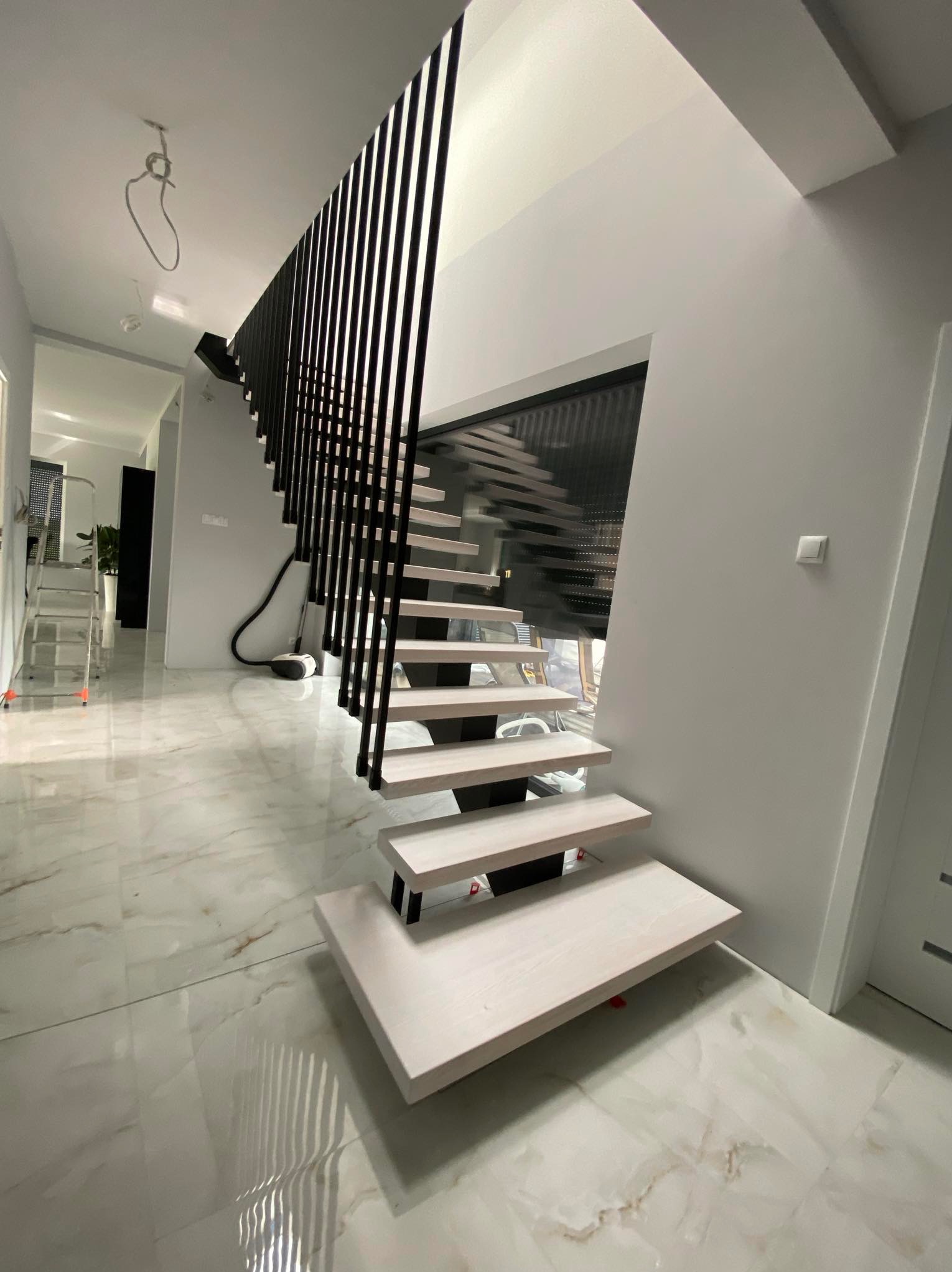 Nowoczesne schody z drewnianymi stopniami i czarną, pionową balustradą, widoczne na tle marmurowej podłogi, z elementami instalacji elektrycznej na suficie w tle.