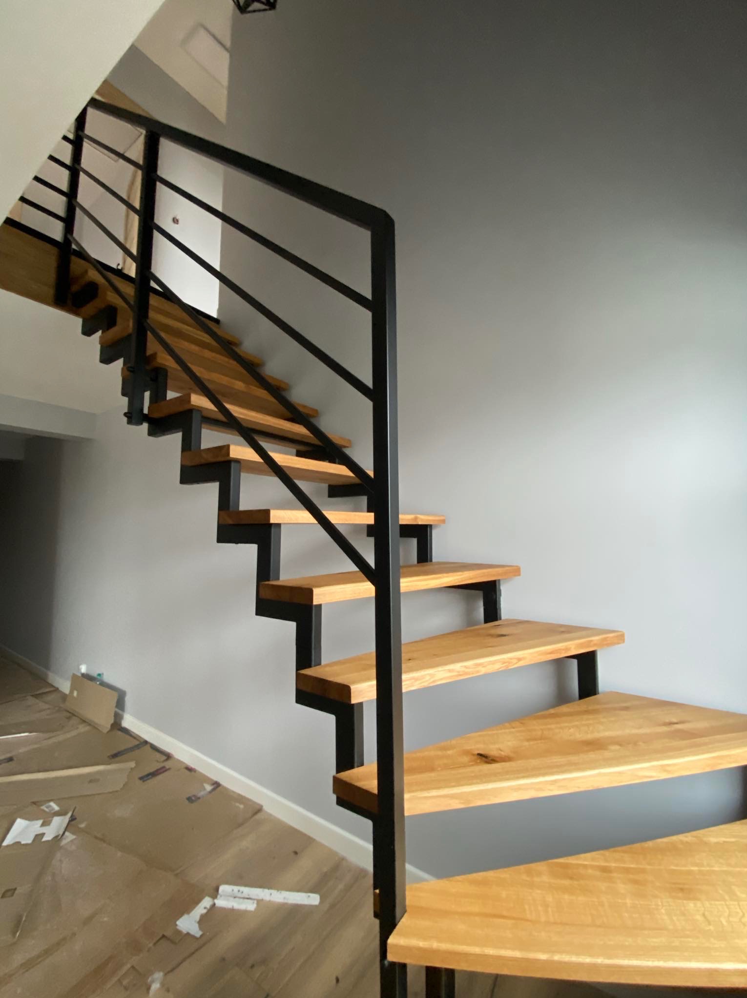 Ażurowe schody z drewnianymi stopniami i czarną, metalową balustradą w nowoczesnym wnętrzu podczas montażu, widoczne kartony ochronne na podłodze.