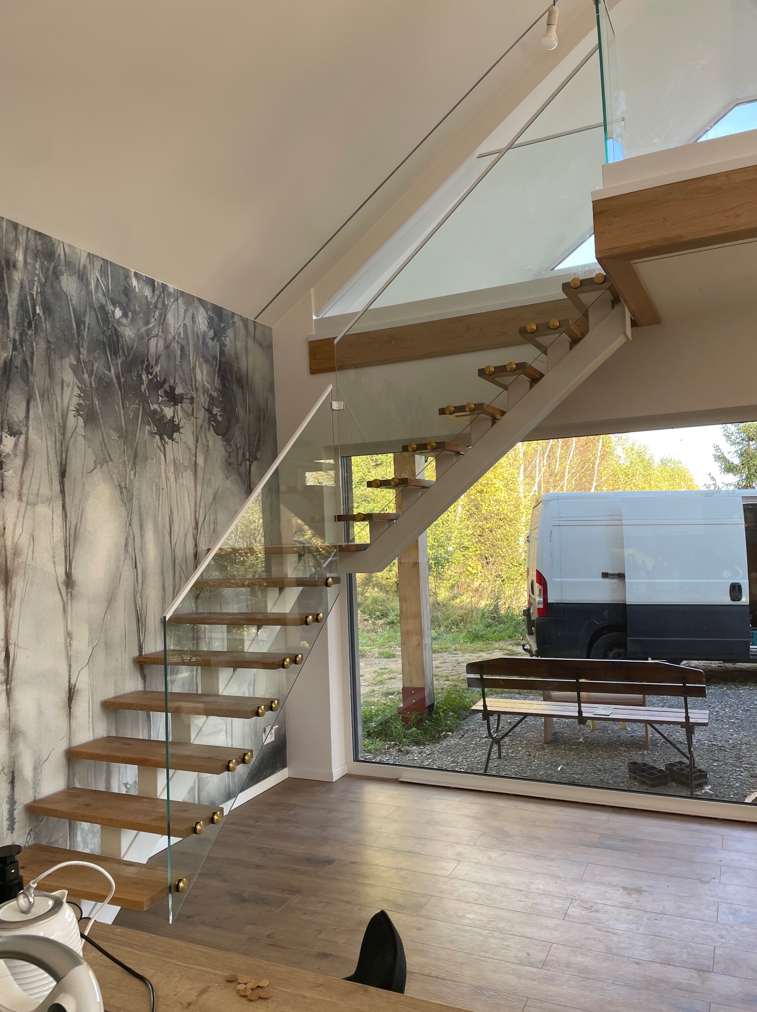 Nowoczesne schody z drewnianymi stopniami i szklaną balustradą w jasnym wnętrzu z tapetą w motywie leśnym, widok na zewnątrz przez duże okno.