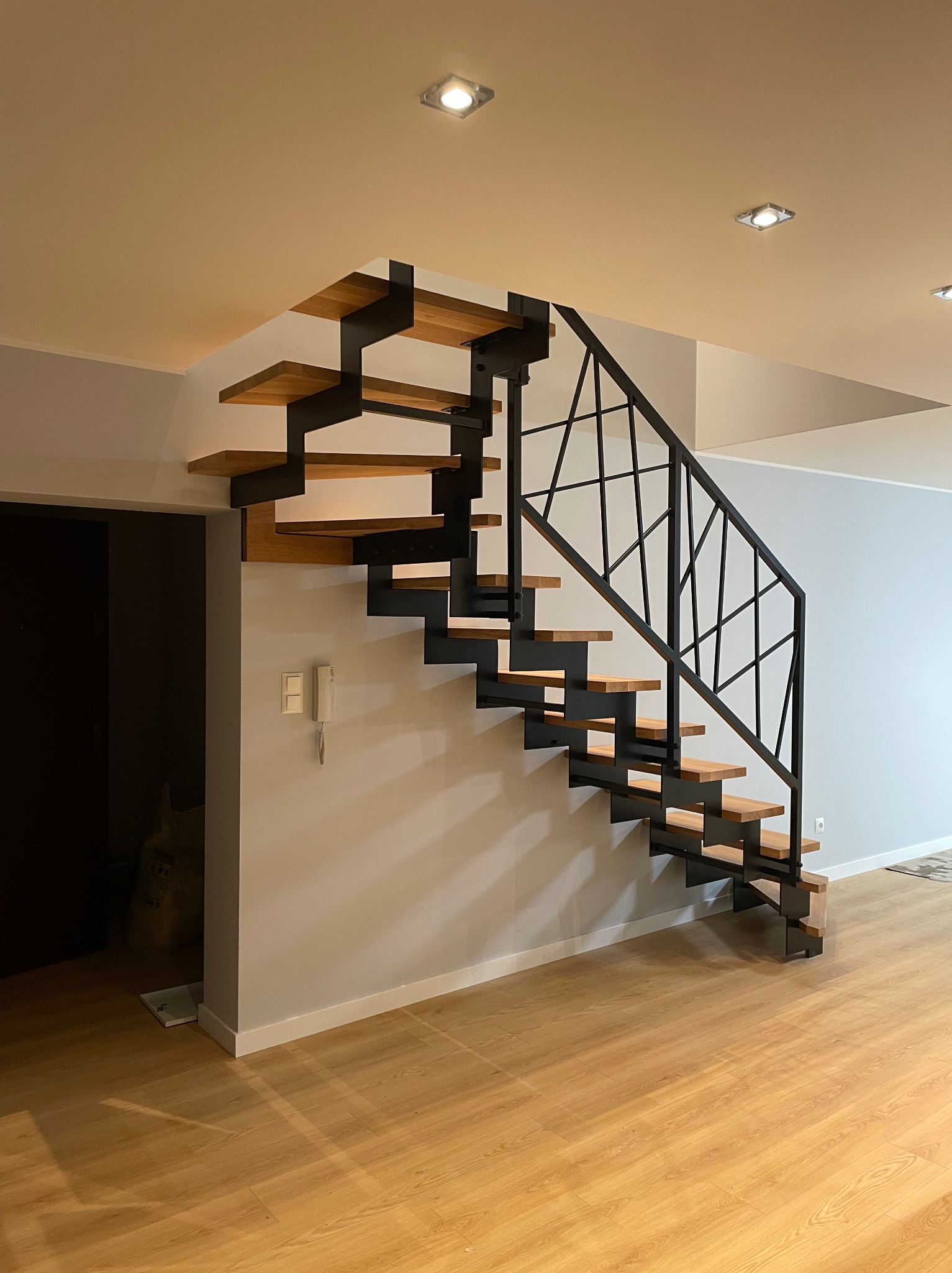 Nowoczesne schody z drewnianymi stopniami i czarną, geometryczną balustradą, widoczne w jasnym wnętrzu z drewnianą podłogą. Minimalistyczny design.