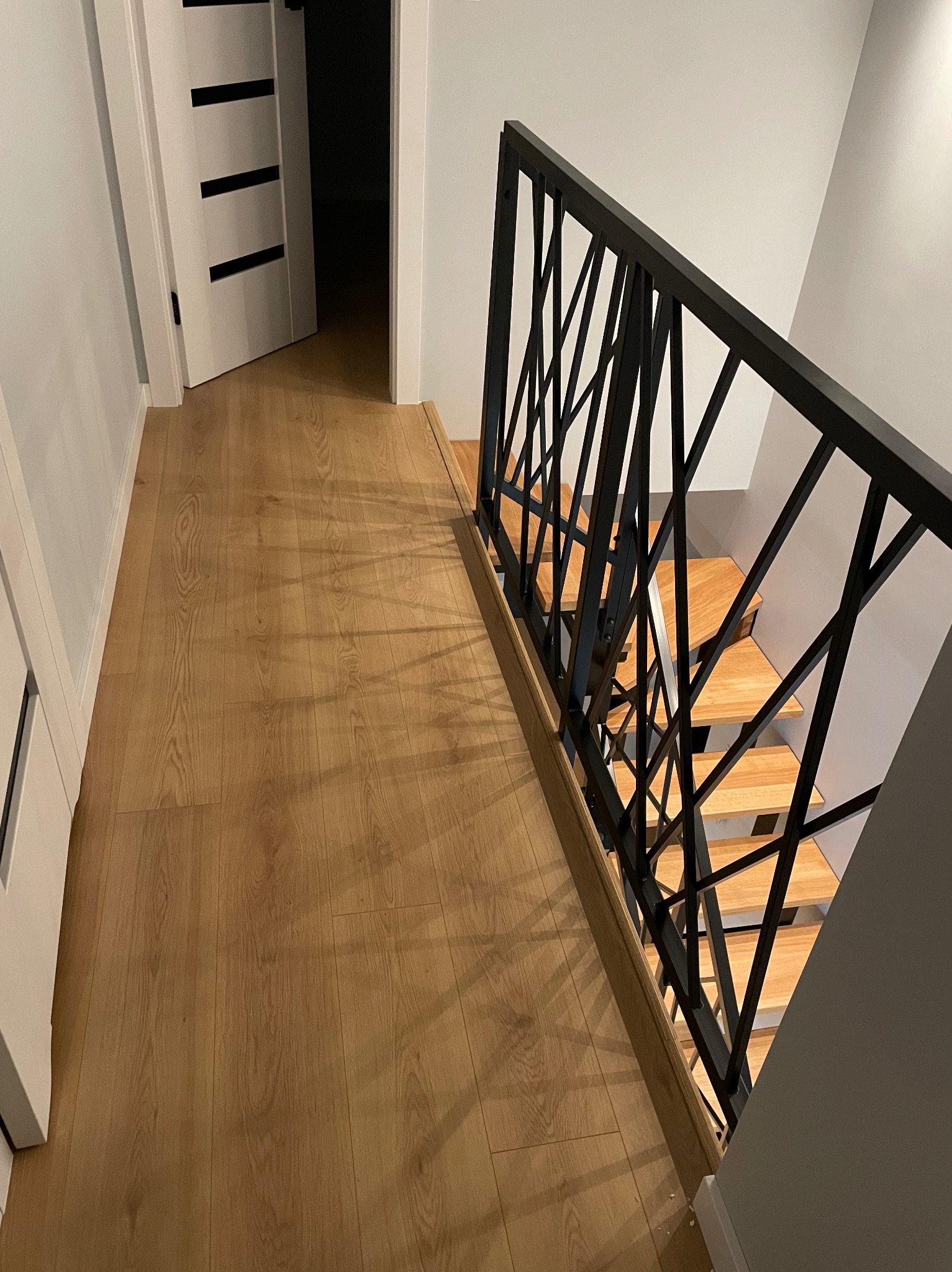 Nowoczesne schody z drewnianymi stopniami i czarną metalową balustradą o geometrycznym wzorze, widoczne z góry, w jasnym wnętrzu z drewnianą podłogą i białymi drzwiami.