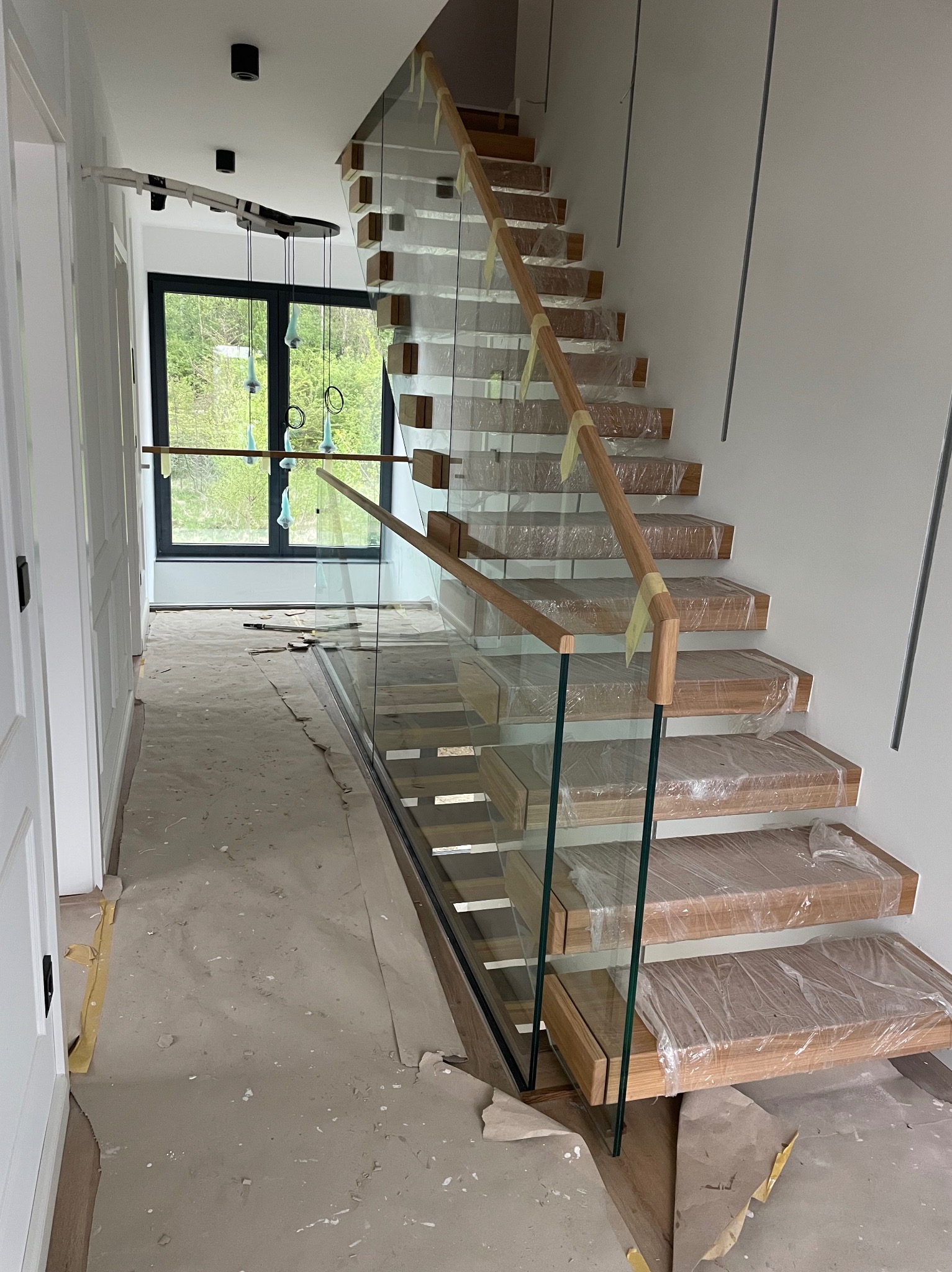 Nowoczesne schody drewniane z balustradą szklaną i drewnianą poręczą, zabezpieczone folią ochronną podczas prac wykończeniowych w jasnym wnętrzu domu.