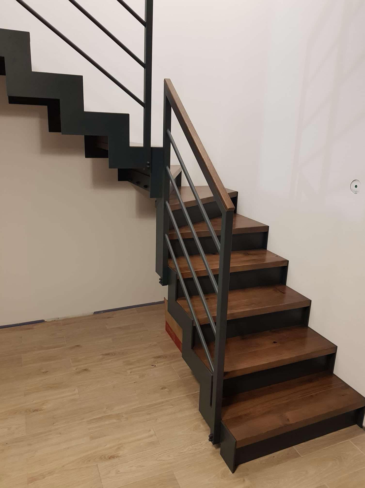 Nowoczesne schody z drewnianymi stopniami i metalową balustradą, widoczne od dołu, z drewnianą poręczą, na tle białej ściany i podłogi z płytek imitujących drewno.