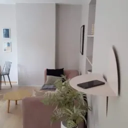 Jasne wnętrze pokoju dziennego z drewnianą podłogą, okrągłym stołem z krzesłami, różową sofą i półką ścienną z telefonem.