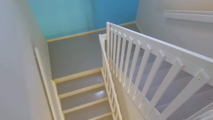 Świeżo pomalowane schody z białymi drewnianymi balustradami, szare ściany i błękitny sufit w górnej części klatki schodowej.