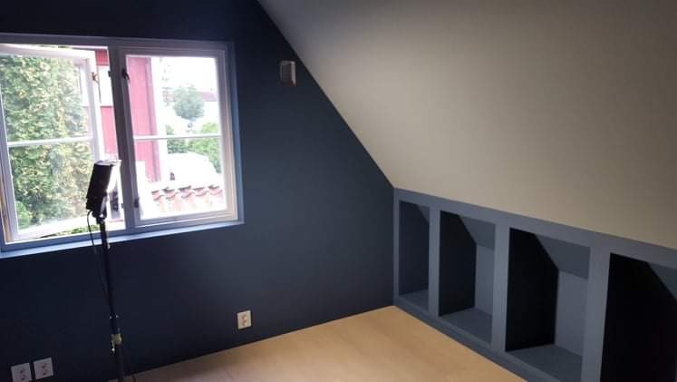 Poddasze po remoncie: ściana pomalowana na granatowo, wnęki w ścianie z szarymi obramowaniami, otwarte okno z widokiem na zieleń, mikrofon na statywie.