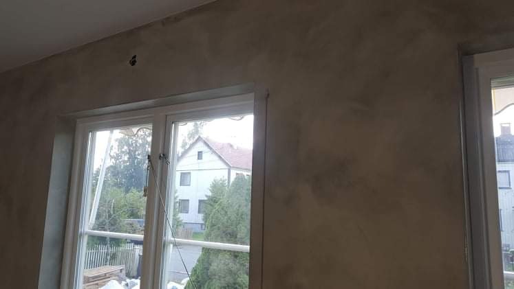 Ściana wewnętrzna z efektem dekoracyjnym, widoczne okno z widokiem na dom i zieleń, jasne ramy okienne, nieregularna faktura na ścianie.