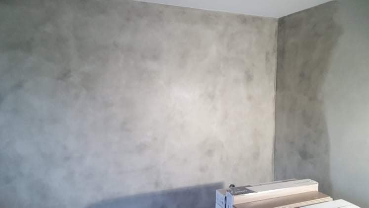 Ściana wewnętrzna pokryta dekoracyjną szpachlą w odcieniach szarości, imitująca efekt betonu architektonicznego, z widocznym stosem paneli podłogowych u dołu.