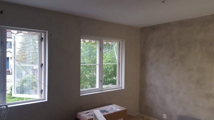 Ściana w pokoju po szpachlowaniu i malowaniu na szaro, z widocznymi dwoma oknami z białymi ramami i kartonami z narzędziami na podłodze.