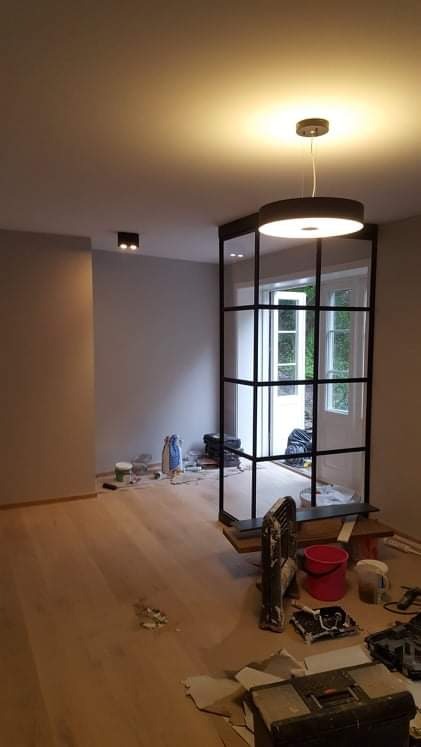 Wnętrze w trakcie prac wykończeniowych: drewniana podłoga, szklana ścianka działowa z czarnymi szprosami, widoczne narzędzia i materiały budowlane, dwa rodzaje oświetlenia sufitowego.