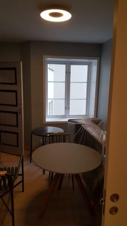 Wnętrze pokoju w trakcie remontu: meble przykryte folią ochronną, okrągły stół z drewnianymi nogami na pierwszym planie, okno z widokiem na budynek naprzeciwko, okrągła lampa sufitowa.