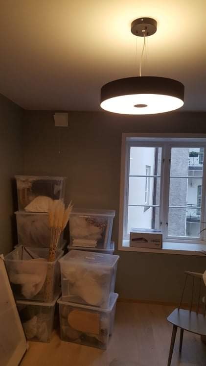 Pokój w trakcie przeprowadzki lub remontu, z plastikowymi pojemnikami wypełnionymi rzeczami, okrągłą lampą sufitową i krzesłem w tle okna.