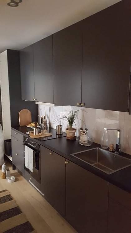 Nowoczesna kuchnia z ciemnymi, matowymi szafkami, marmurowym backsplashiem, drewnianą deską do krojenia i metalowymi akcesoriami na blacie, oświetlona naturalnym światłem.