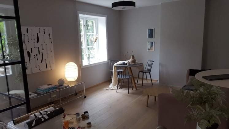 Jasne, minimalistyczne wnętrze po remoncie: drewniana podłoga, szare ściany, okno z widokiem na zieleń, okrągły stół z krzesłami, designerska lampa i dekoracje.