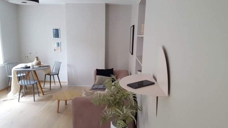 Jasne wnętrze pokoju dziennego z drewnianą podłogą, okrągłym stołem z krzesłami, różową sofą i półką ścienną z telefonem.