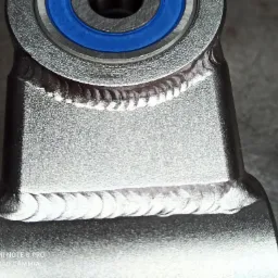 Szlifowany metalowy element konstrukcyjny z widocznymi spawami i niebieskim łożyskiem, sfotografowany z bliska.