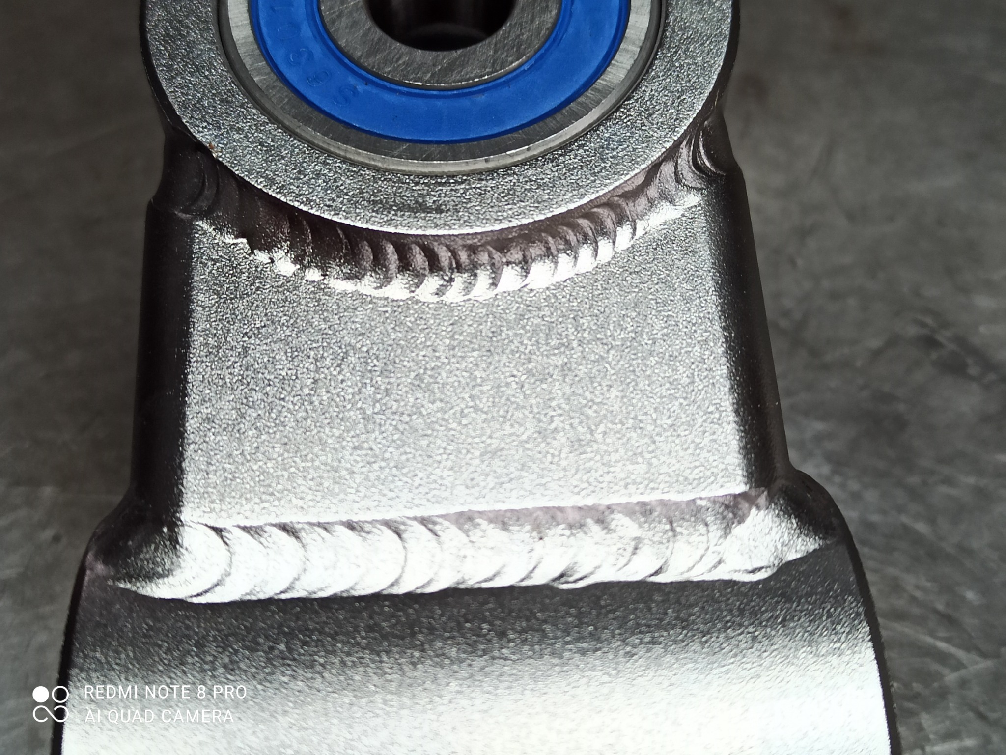 Szlifowany metalowy element konstrukcyjny z widocznymi spawami i niebieskim łożyskiem, sfotografowany z bliska.