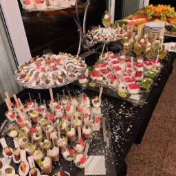 Bogato zastawiony stół cateringowy z deserami: mini-pucharki z kremem i owocami, galaretki, czekoladowe trufle na łyżeczkach i patery z owocami, dekorowany złotymi konfetti w kształcie gwiazdek.