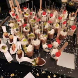 Deserki w małych plastikowych kubeczkach i na łyżeczkach, z bitą śmietaną, owocami (wiśnie, kiwi, borówki), czekoladą, ułożone na czarnym stole posypanym białym i złotym konfetti.