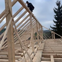 Dachroof - Konstrukcja drewniana dachu w trakcie budowy, widoczny dekarz w czapce na szczycie, w tle drzewo iglaste i pochmurne niebo.