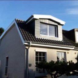 Dachroof - Dwukondygnacyjny dom z szarej cegły z ciemnym dachem i oknami w lukarnach pod błękitnym niebem.