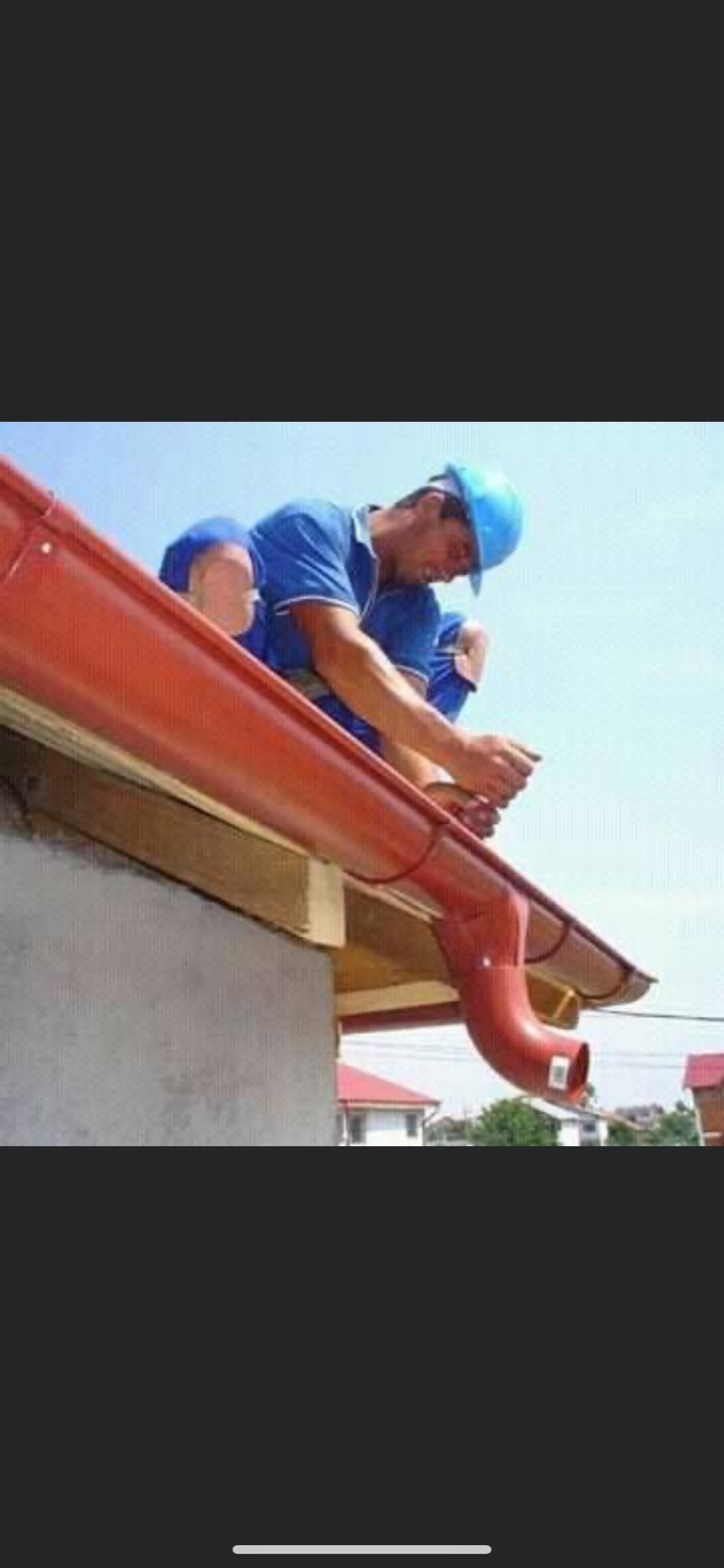 Monter w niebieskim kasku i ubraniu instaluje czerwoną rynnę dachową, widoczny fragment dachu i budynki w tle.