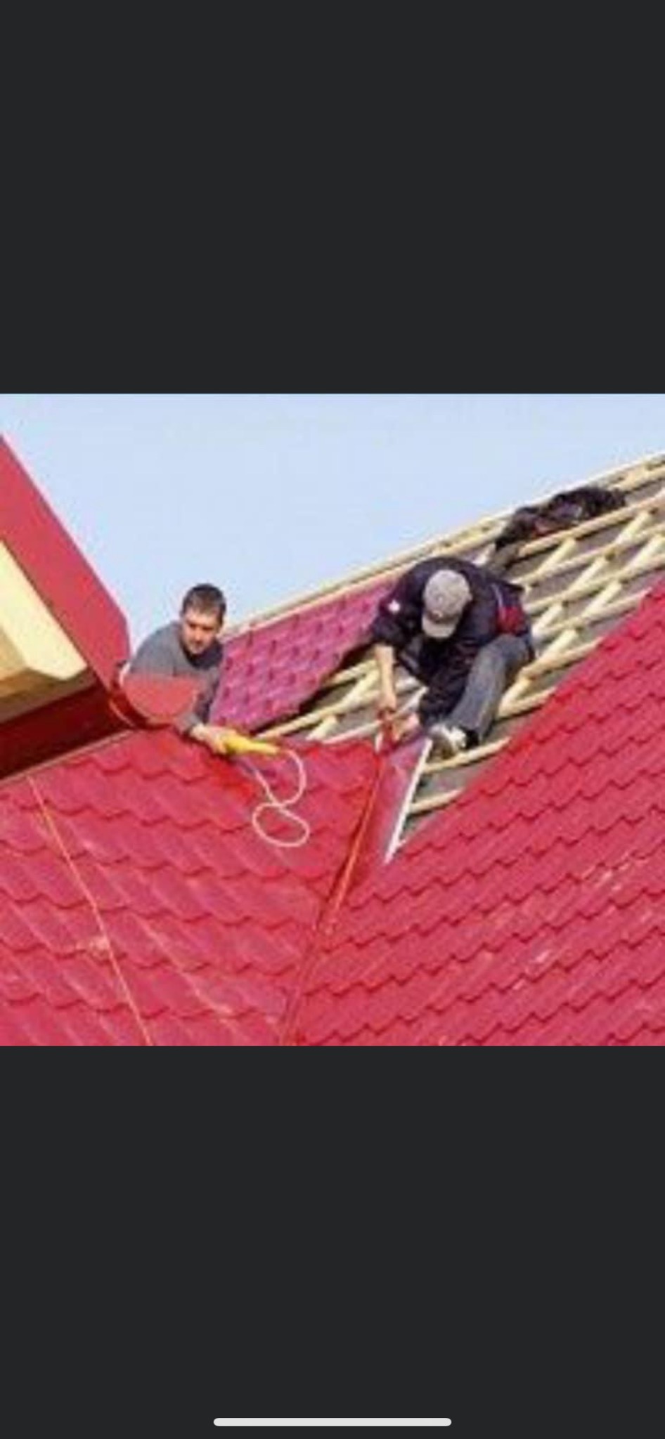 Dwóch dekarzy pracuje na dachu pokrytym czerwoną dachówką, jeden uszczelnia połączenie, drugi podaje kolejne elementy pokrycia.