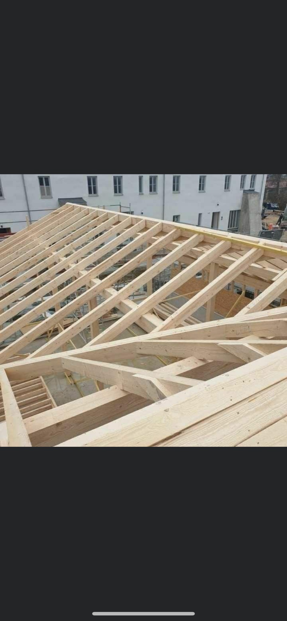 Surowa drewniana konstrukcja dachu z widocznymi krokwiami i łatami, na tle budynku z oknami i kominem.