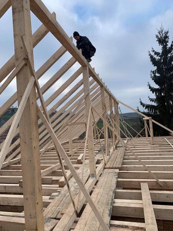 Konstrukcja drewniana dachu w trakcie budowy, widoczny dekarz w czapce na szczycie, w tle drzewo iglaste i pochmurne niebo.