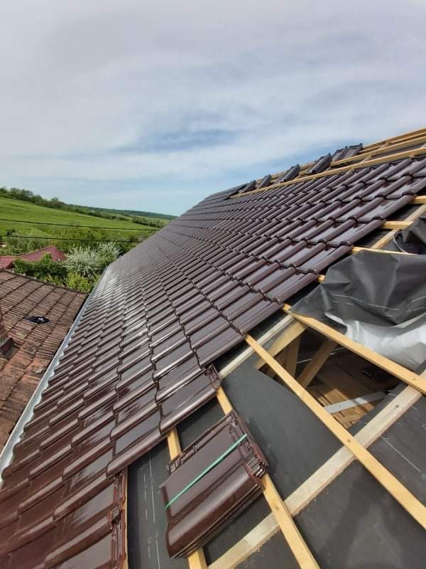 Układanie brązowej dachówki ceramicznej na dachu z widoczną konstrukcją drewnianą i membraną dachową, w tle zielony krajobraz.