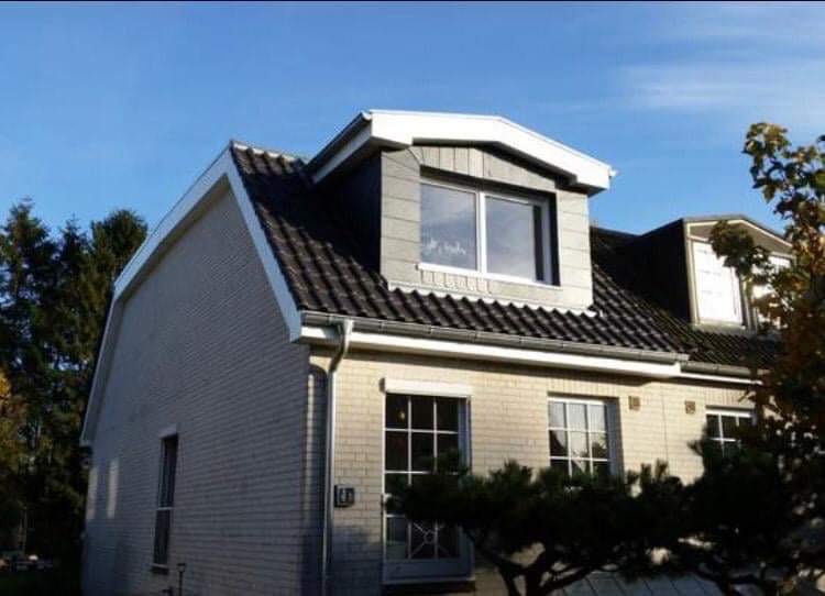 Dwukondygnacyjny dom z szarej cegły z ciemnym dachem i oknami w lukarnach pod błękitnym niebem.