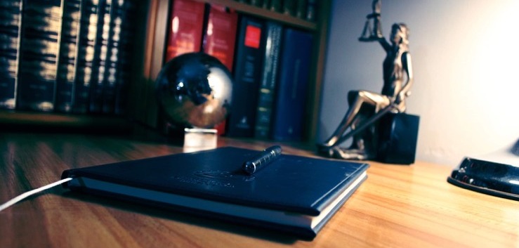 Biurko z notatnikiem, długopisem i figurką Temidy na tle regału z książkami. Nastrojowe, stonowane oświetlenie.