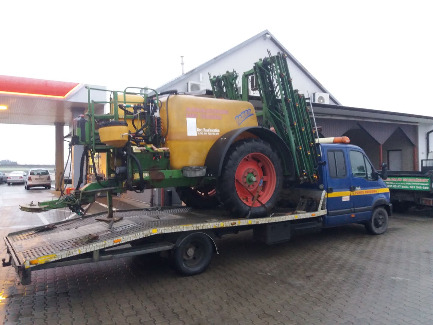 Niebieska laweta przewozi żółty opryskiwacz rolniczy z czerwonymi felgami na tle stacji benzynowej.