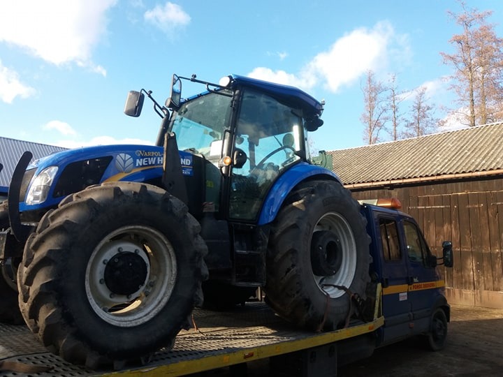 Niebieski traktor New Holland przewożony na platformie małej, granatowej ciężarówki z żółtymi pasami, na tle budynku gospodarczego i błękitnego nieba.