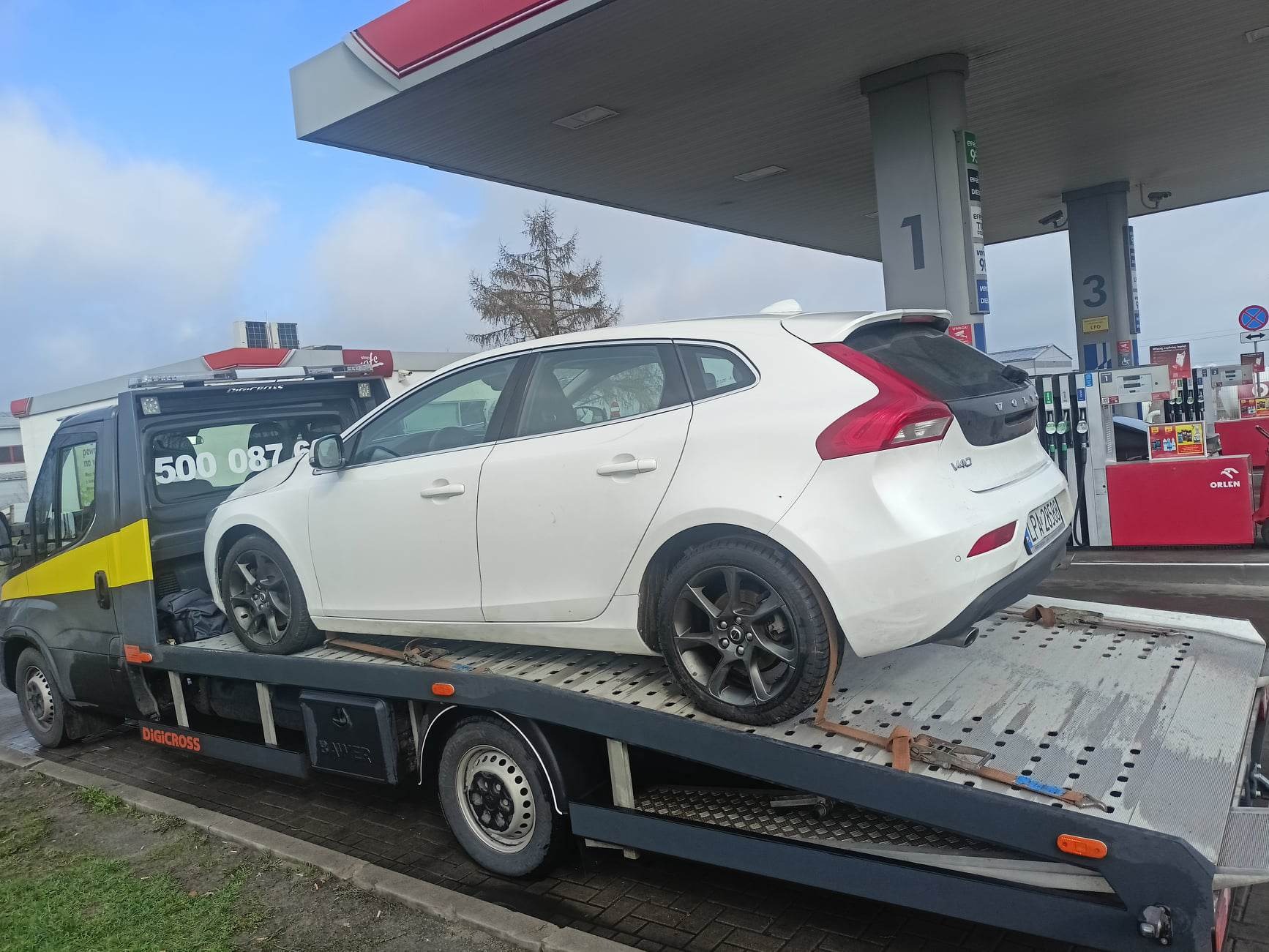 Biały samochód osobowy Volvo V40 zabezpieczony pasami na platformie lawety, na tle stacji benzynowej Orlen.