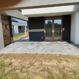 Nowoczesny taras wyłożony szarą kostką brukową o geometrycznym wzorze, przylegający do budynku z ciemnoszarą elewacją i drewnianymi oknami. Na pierwszym planie widoczny pas ziemi z resztkami trawy,...