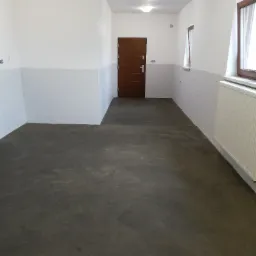 Świeżo wylana, szara posadzka w pomieszczeniu z białymi ścianami i brązowymi drzwiami, widoczny grzejnik pod oknem.