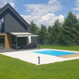 Nowoczesny dom z ciemną elewacją, jasnym fragmentem i drewnianą okładziną, pergola z meblami ogrodowymi i basen na zadbanym trawniku w słoneczny dzień.