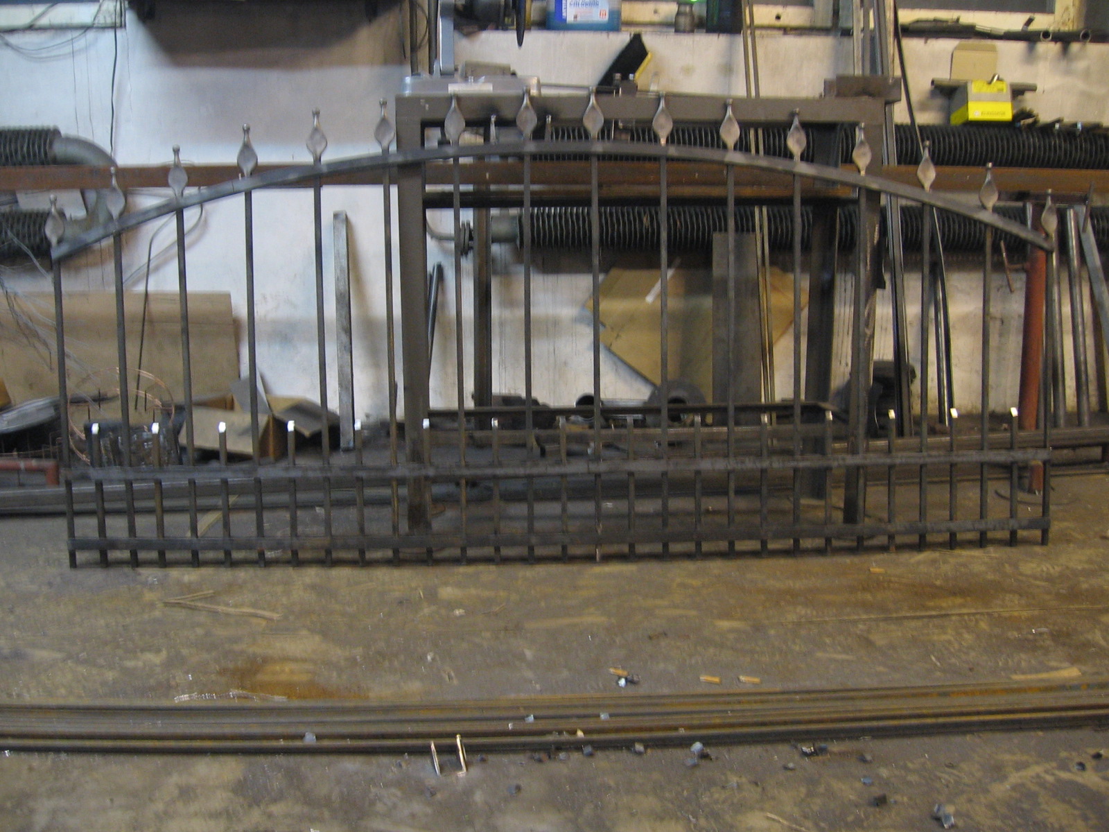 Surowa, metalowa konstrukcja bramy z ozdobnymi grotami, leżąca na podłodze warsztatu w trakcie produkcji, z widocznymi elementami wyposażenia warsztatowego w tle.