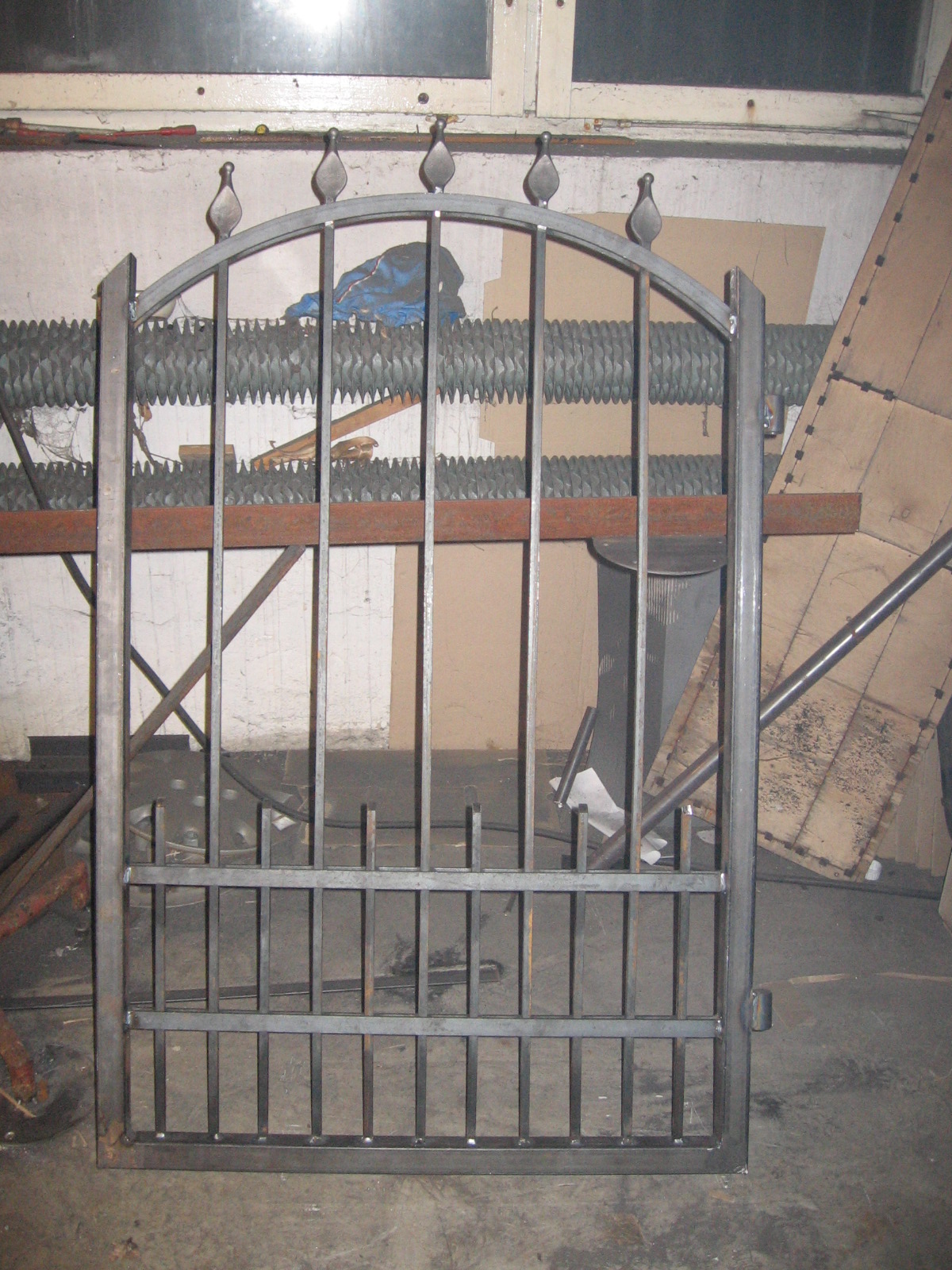 Surowa, metalowa brama ogrodzeniowa z łukiem i ozdobnymi grotami, w warsztacie, widoczne narzędzia i nieporządek w tle.