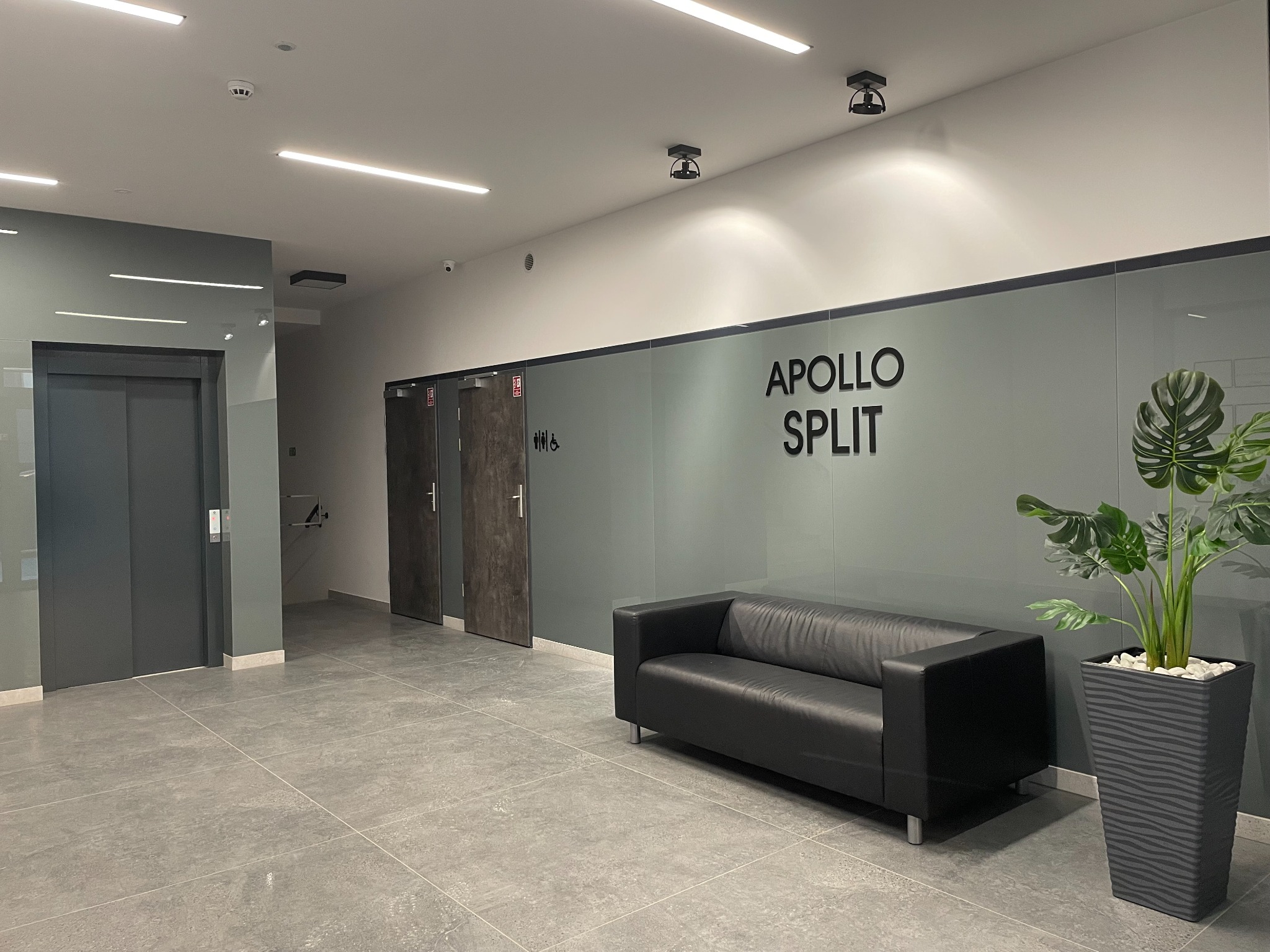Nowoczesne wnętrze poczekalni z czarną skórzaną sofą, rośliną w donicy, szarą podłogą i napisem 'Apollo Split' na szklanej ścianie.