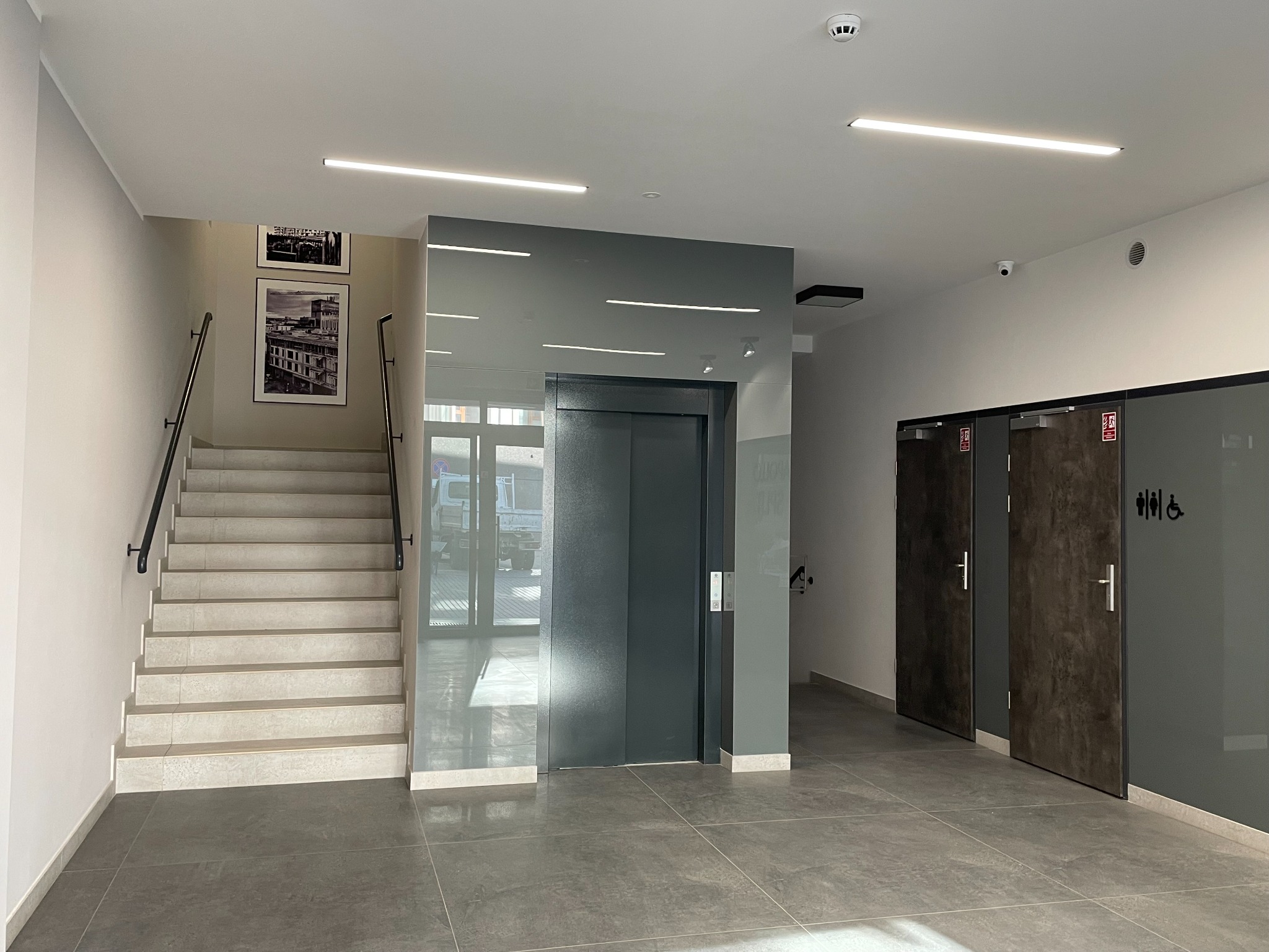 Wnętrze budynku z klatką schodową, windą i toaletami, oświetlone liniowymi lampami LED, z dekoracyjnymi obrazami na ścianie.