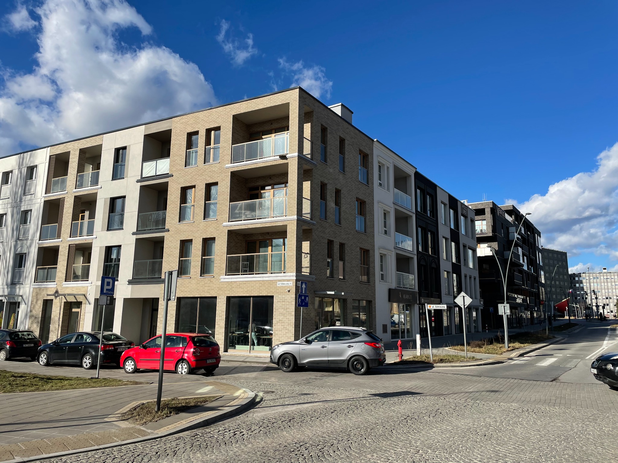 Nowoczesny budynek mieszkalny z balkonami, widoczny z poziomu ulicy wyłożonej kostką brukową, z zaparkowanymi samochodami i znakami drogowymi w słoneczny dzień.