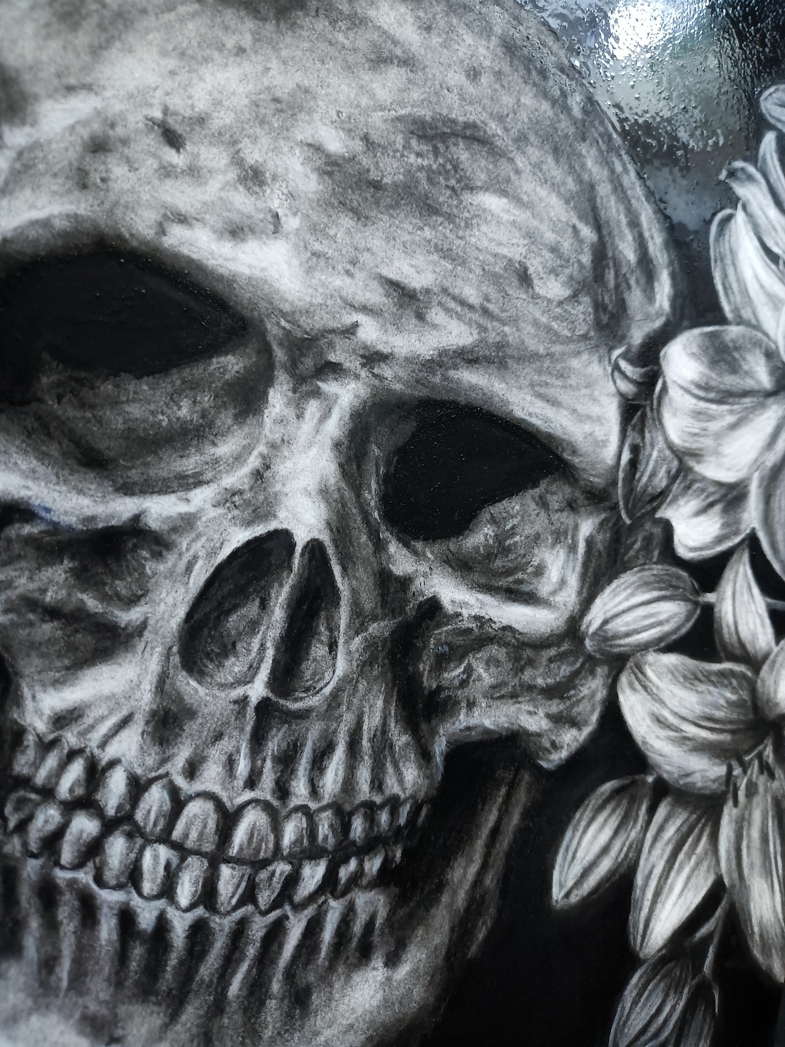 Czarno-biały rysunek węglem przedstawiający czaszkę z kwiatami, wykonany na błyszczącej powierzchni, widoczne drobne refleksy światła.